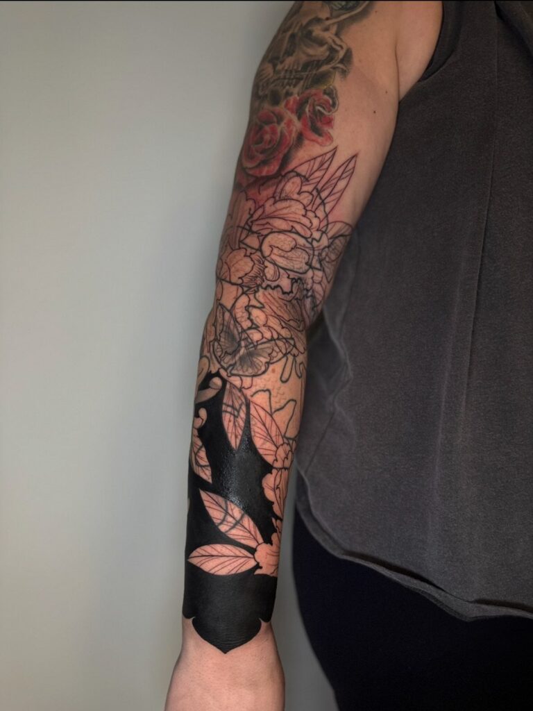 Tattooed arm showing a floral sleeve with a heavy black ink cover up section.