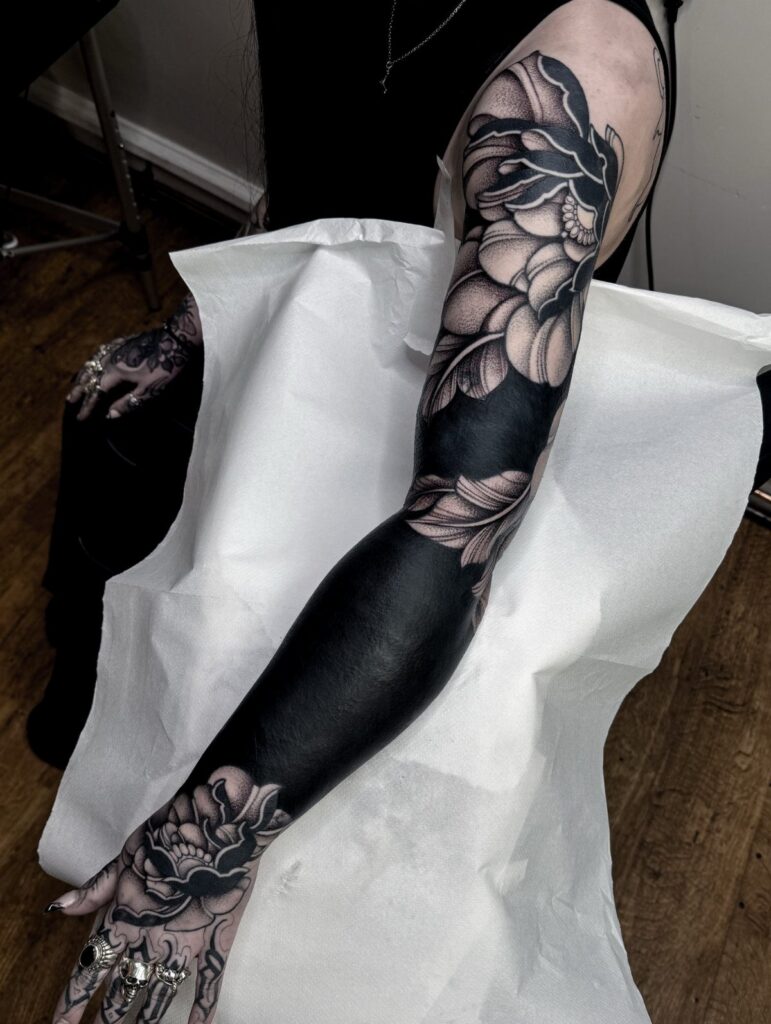 Blackout tattoo cover-up on an arm with grey floral designs on shoulder and hand