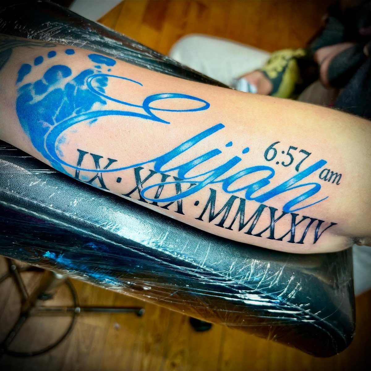 Blue ink tattoo on a forearm with a footprint, name, and Roman numerals.
