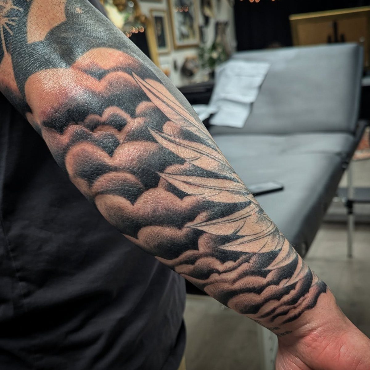 Forearm tattoo featuring black and grey stylized clouds and wind swirls filling the arm.