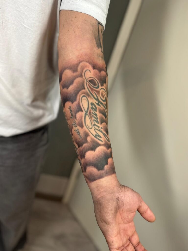 Forearm tattoo featuring grey shaded clouds filling the background around cursive text.