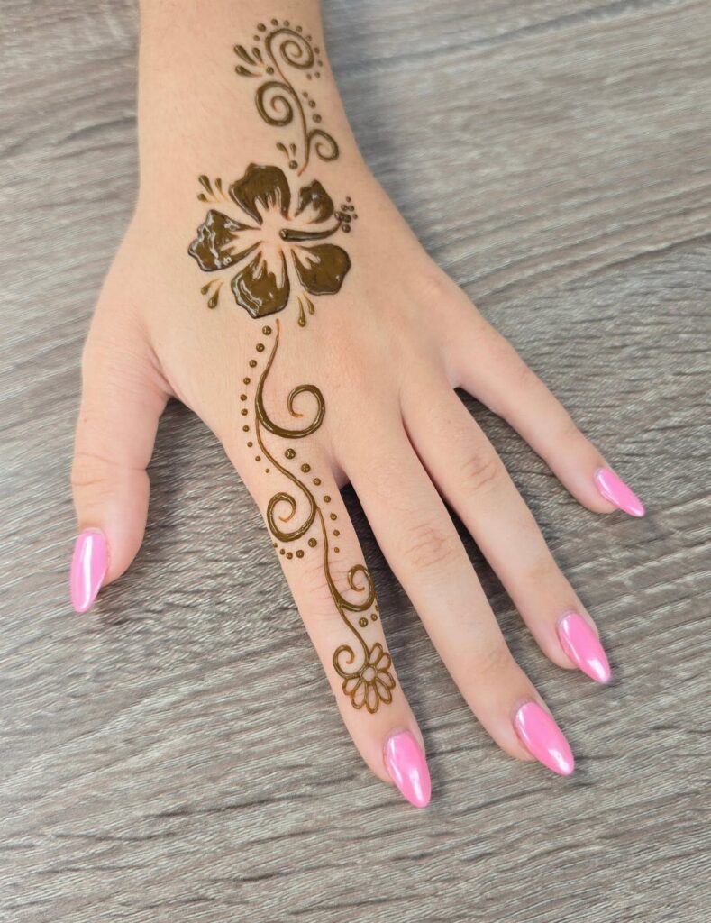 Hand with pink nails featuring a brown henna hibiscus flower and swirl design.
