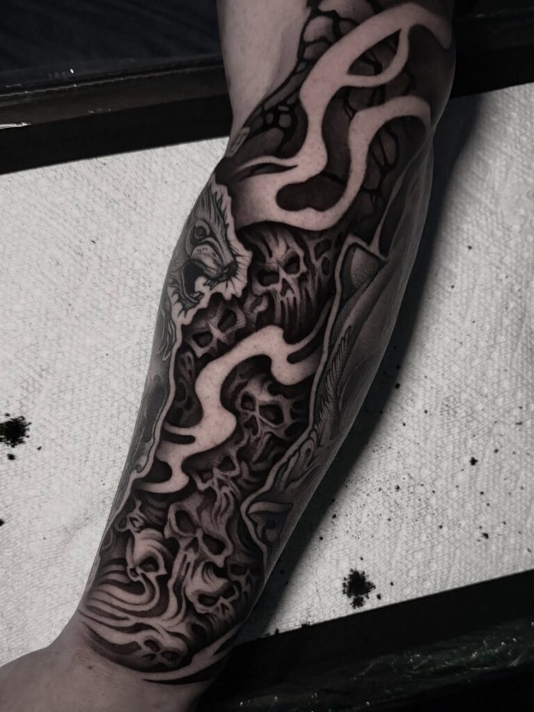 Forearm tattoo featuring black and grey skulls with flowing organic filler patterns.