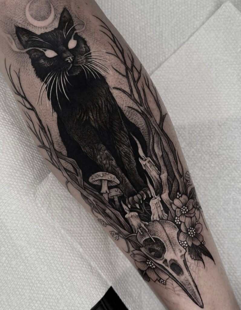 Gothic tattoo of a black cat, bird skull, and crescent moon on an arm.