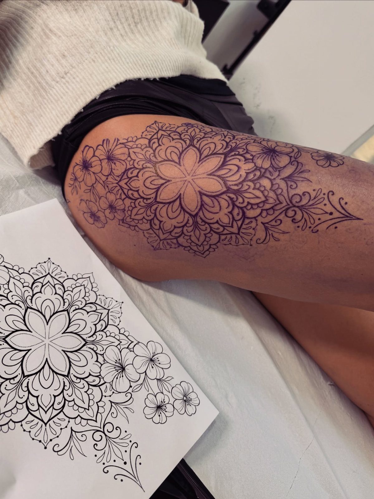 Mandala flower tattoo on a thigh next to its original line drawing stencil.