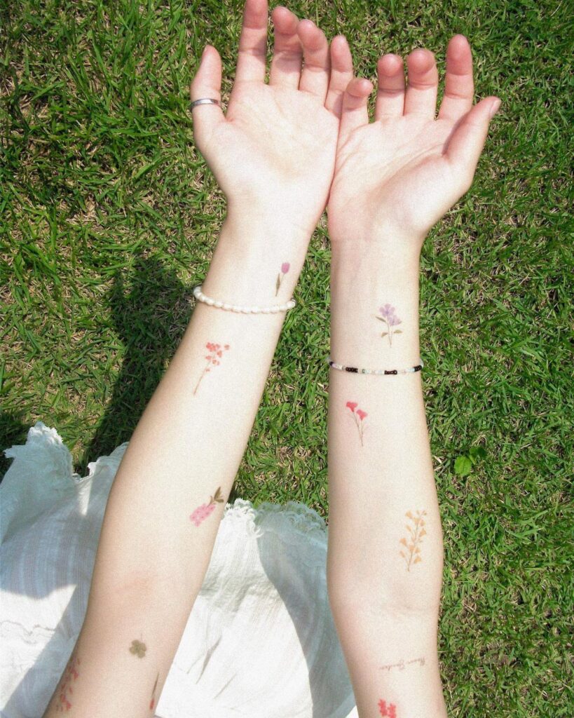 Two forearms with various small floral temporary tattoos resting on green grass.