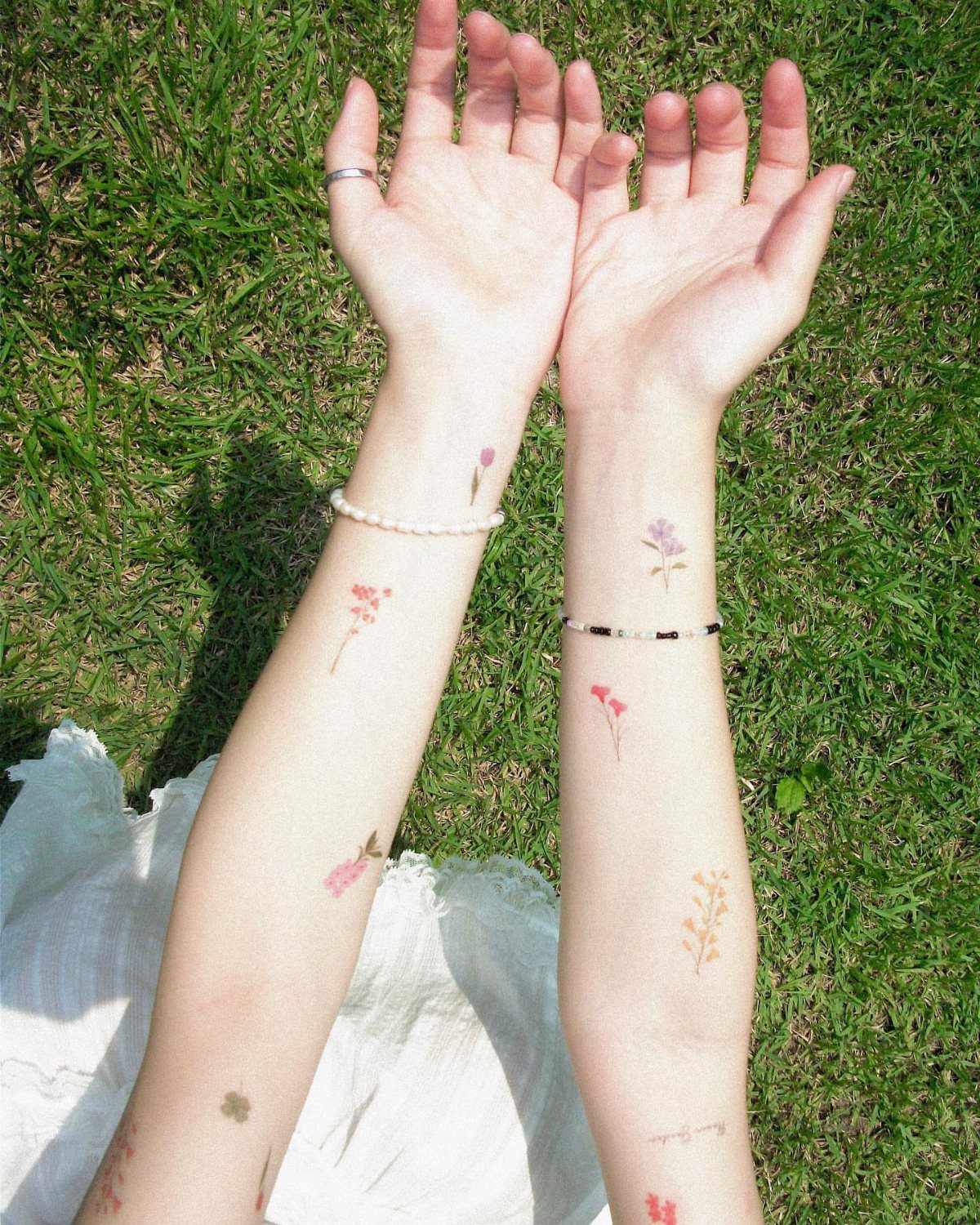 Two forearms with various small floral temporary tattoos resting on green grass.