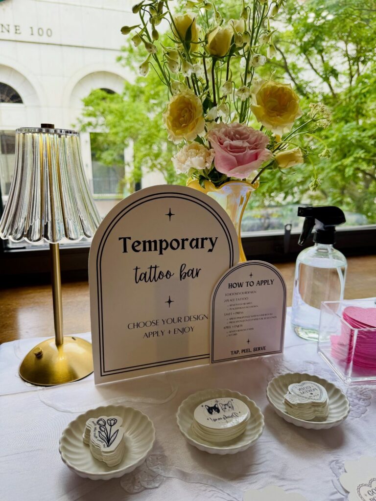 Temporary tattoo bar setup with design stacks, a sign, and fresh flowers.