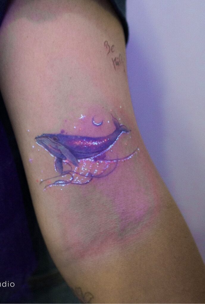 Glowing purple UV tattoo of a whale leaping over a wave on an arm.