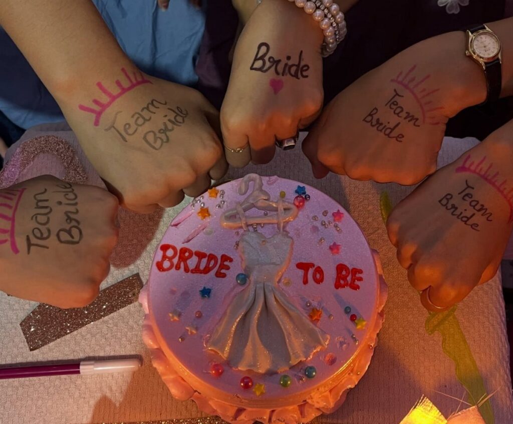 Hands with Team Bride tattoos gathered around a pink Bride to Be cake.