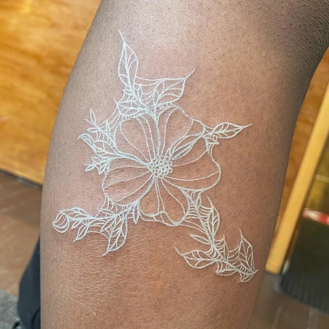 White ink floral tattoo with a flower and leaves on dark brown skin.